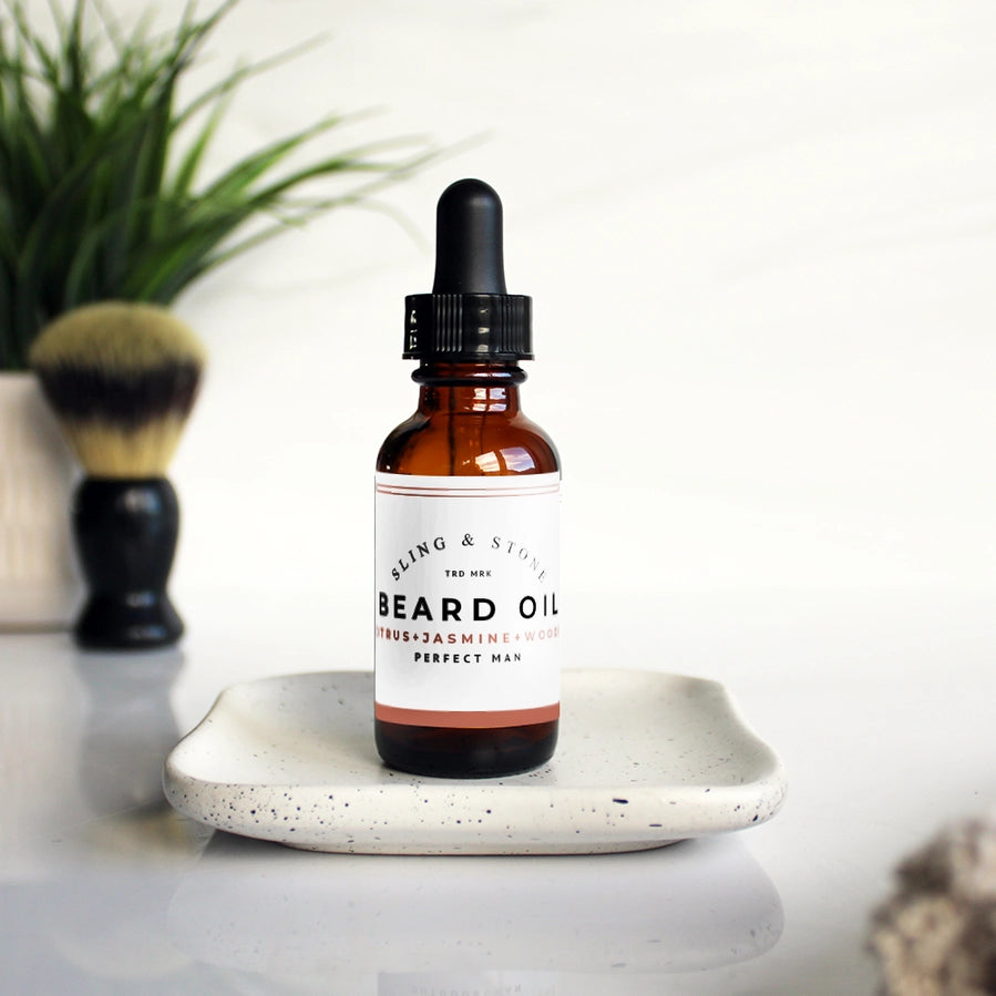 A glass amber bottle of Sling & Stone Beard Oil with a black cap, placed on a ceramic plate with a brush and plant in the background.