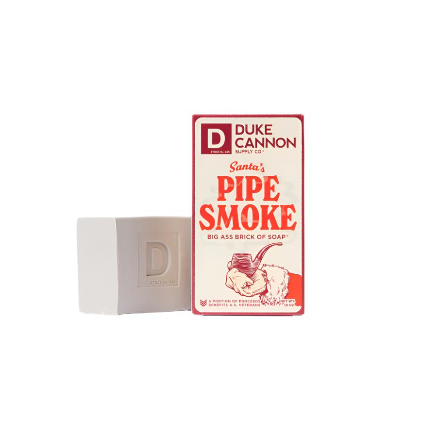 A red and white packaging of Duke Cannon Big Ass Brick of Soap with 'Santa's Pipe Smoke' scent, featuring an illustration of a pipe and Santa Claus.