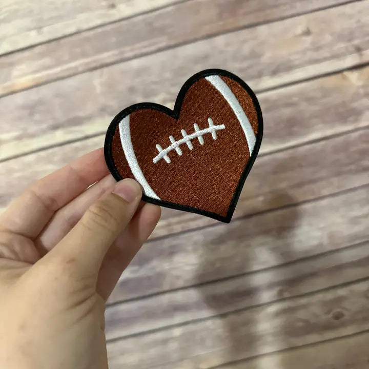 A hand holding a heart-shaped iron-on patch with a football design against a wooden background.