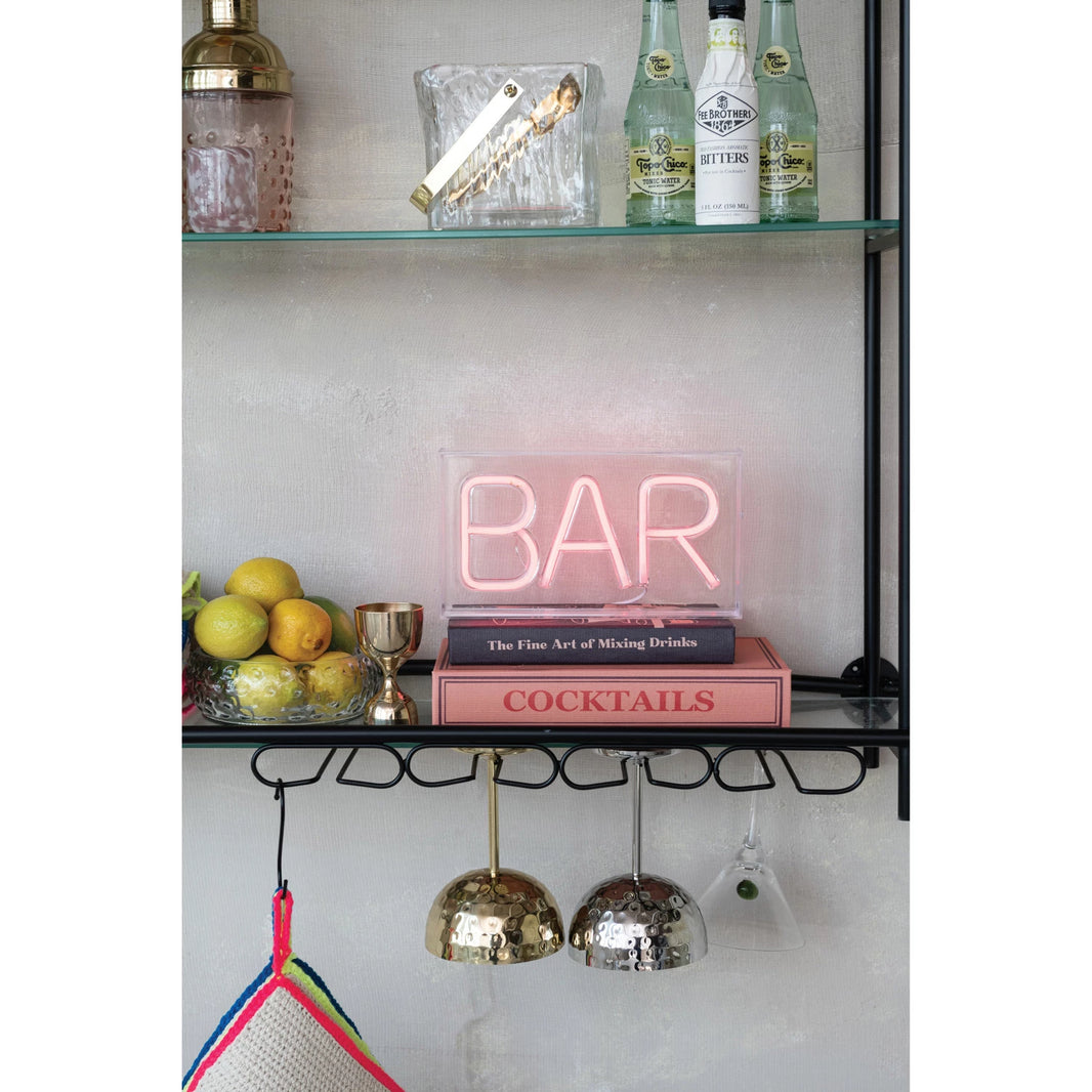 A neon style LED BAR sign sitting on top of books about cocktails on a shelf with other barware.