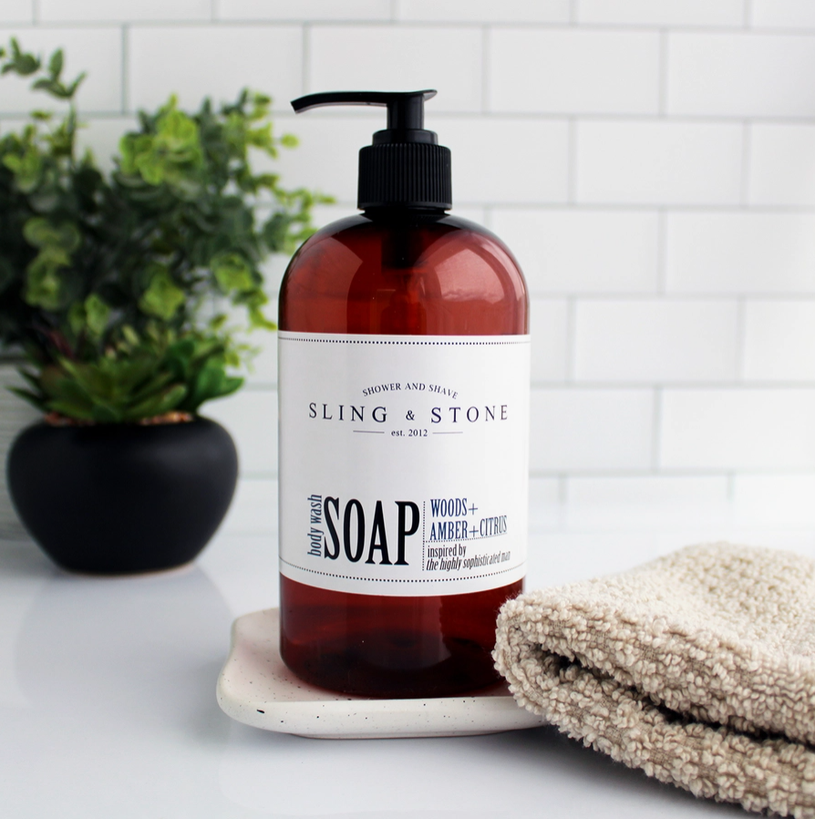 An amber pump bottle labeled 'Sling & Stone Liquid Soap' on a white surface with a beige towel and a plant in the background.
