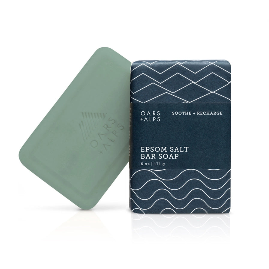 A bar of soap with a greenish tint next to its packaging, which has a blue and white pattern. The packaging reads 'Epsom Salt Bar Soap'.