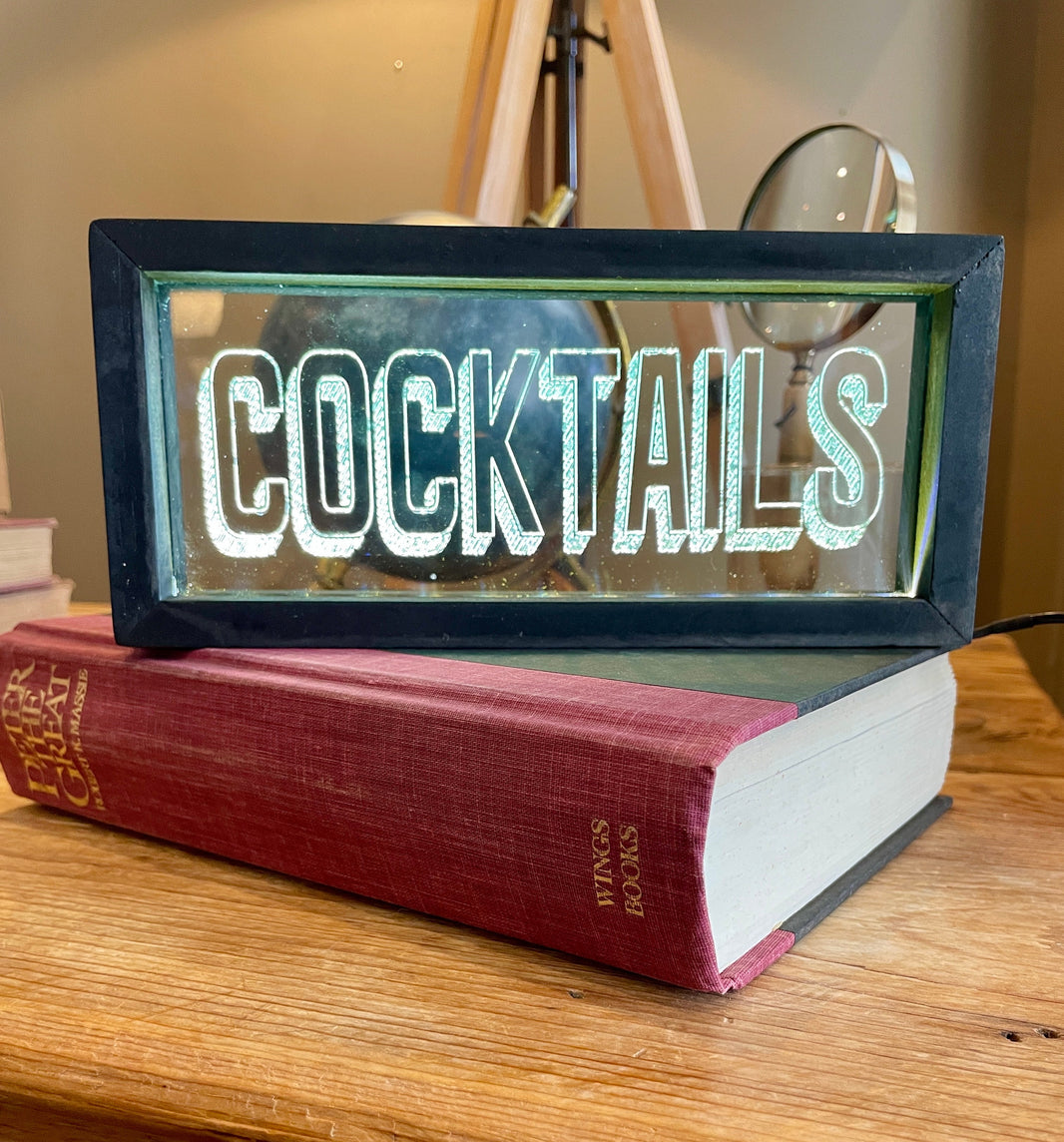 Lighted sign reading 'COCKTAILS' on top of a book on a wooden surface.