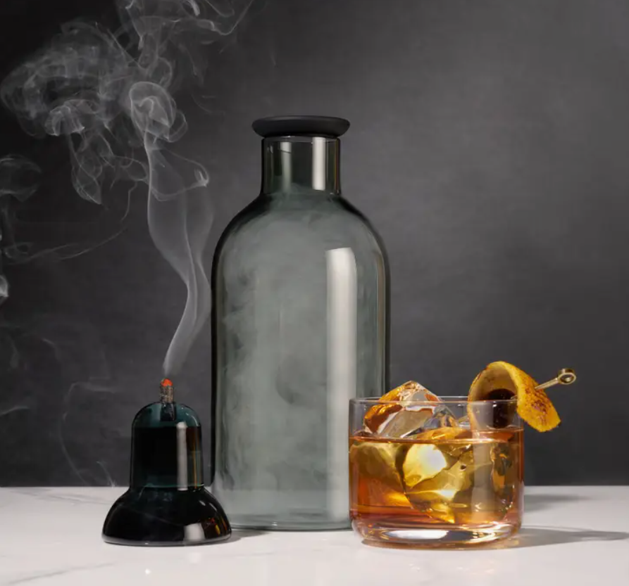 The Viski Smoked Cocktail kit with a lit smoking pellet and the infusion vessel filled with smoke. A glass filled with a cocktail sits nearby.