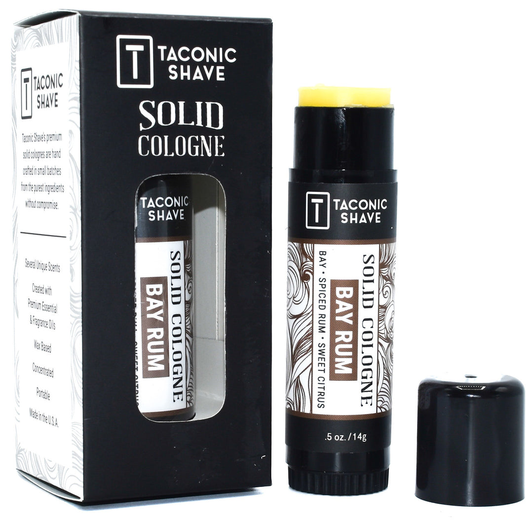 Taconic Shave Bay Rum Solid Cologne, cologne stick, and packaging. The product is displayed in front of its black box packaging with white and brown bay rum themed designs.