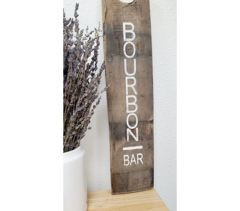 Wooden sign with 'Bourbon Bar' text next to a vase of lavender on a white background