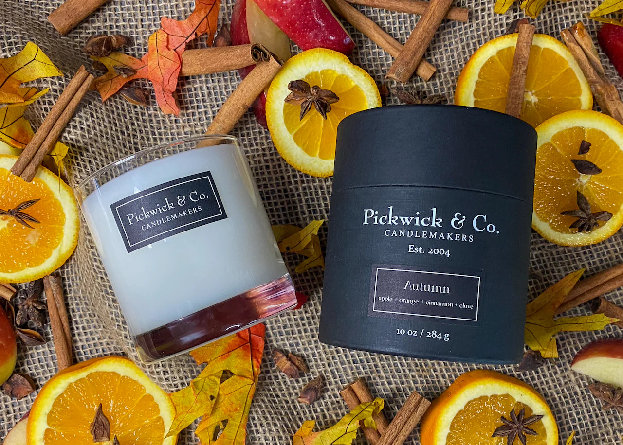 Two candles from Pickwick & Co. Candlemakers surrounded by autumnal elements like oranges, cinnamon sticks, and leaves on a textured surface.