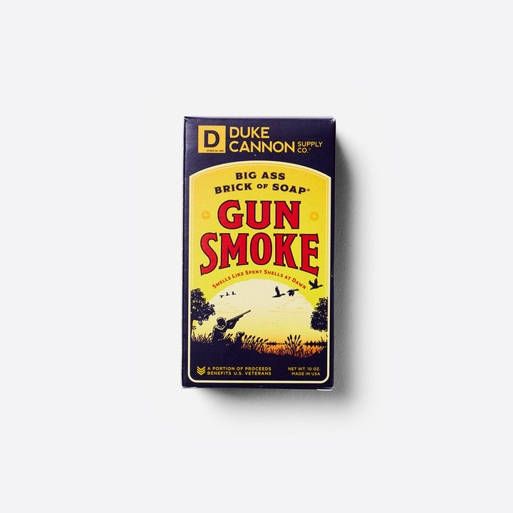 A large bar of soap in its packaging, labeled 'Duke Cannon - Gun Smoke - Big Ass Brick of Soap' with an illustration of a sunset over a field with birds.