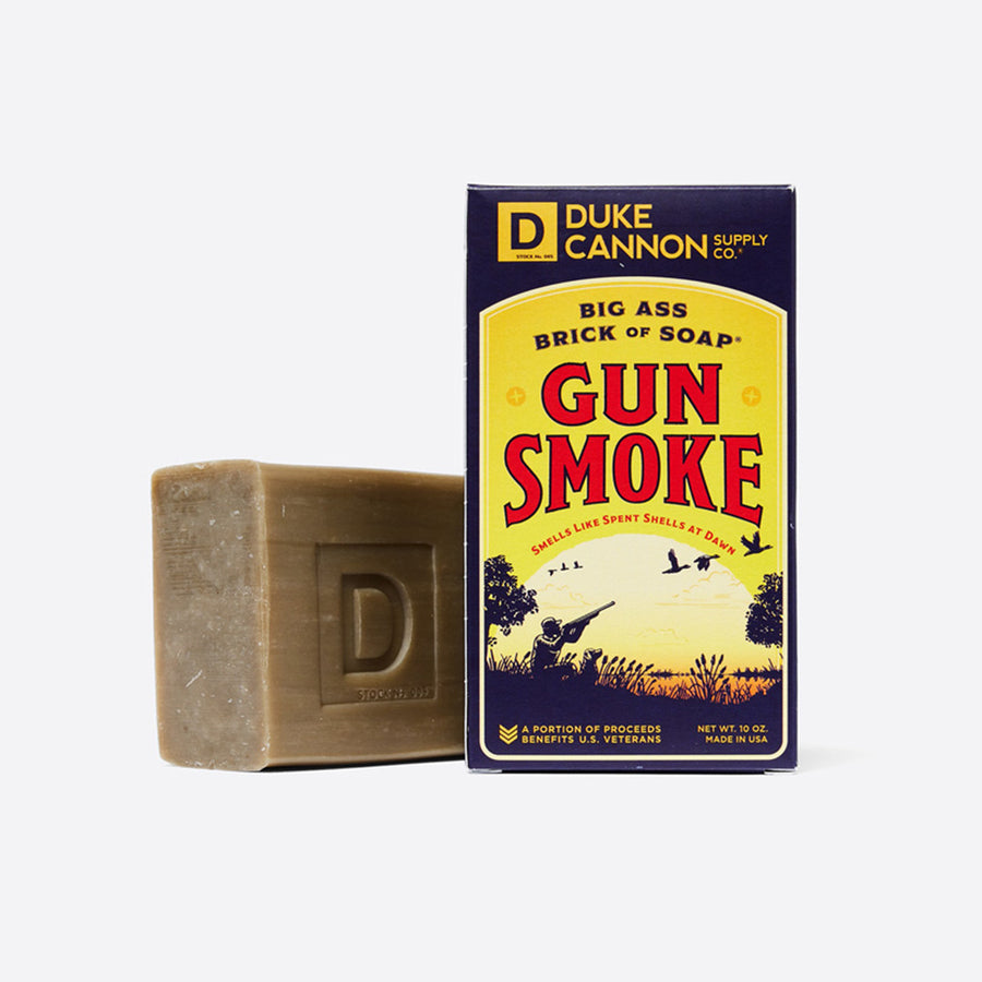 A large bar of soap next to its packaging, labeled 'Duke Cannon - Gun Smoke - Big Ass Brick of Soap' with an illustration of a sunset over a field with birds.