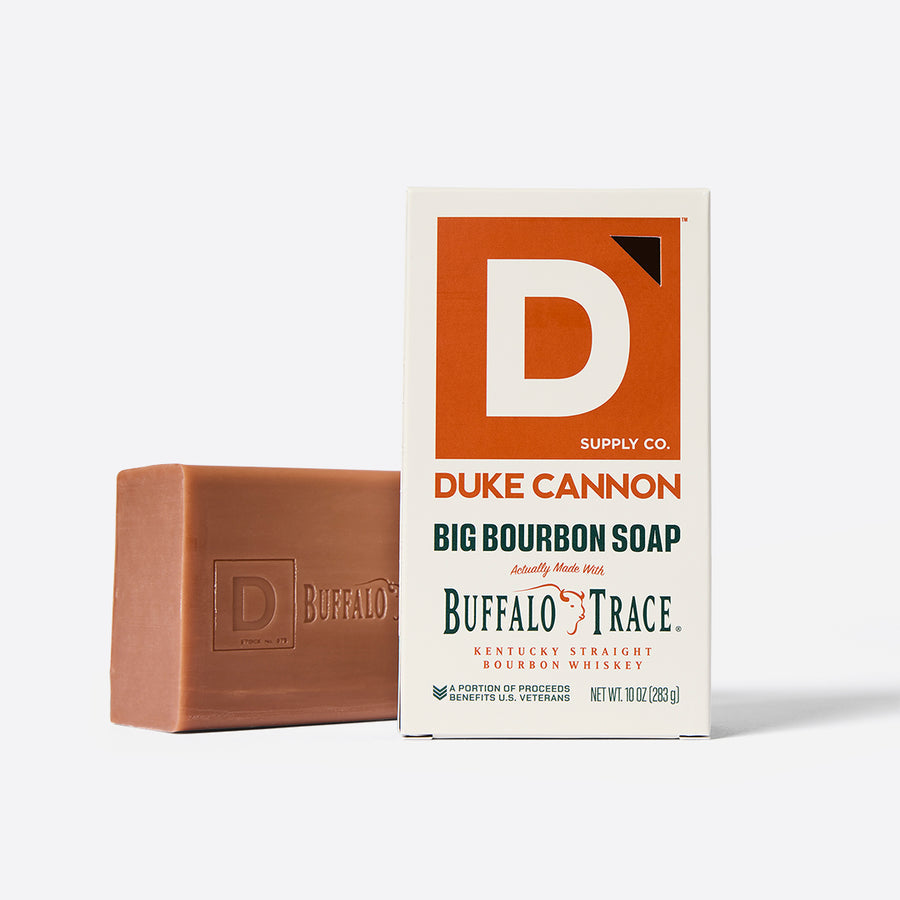 A bar of soap with a brownish tint next to its packaging, which features the Duke Cannon Supply Co. logo and text indicating it is a big bourbon soap with bourbon from Buffalo Trace Distillery.