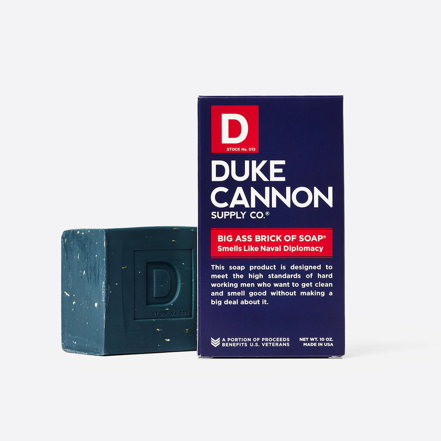 A navy blue bar of soap with exfoliating grains visible on the surface, next to its packaging which reads 'DUKE CANNON SUPPLY CO. BIG ASS BRICK OF SOAP SMELLS LIKE NAVAL DIPLOMACY'.