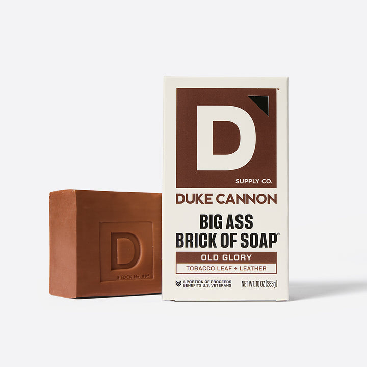A large bar of soap with a brownish-red tint and the Duke Cannon logo embossed on it, next to its packaging that reads 'BIG ASS BRICK OF SOAP OLD GLORY'.
