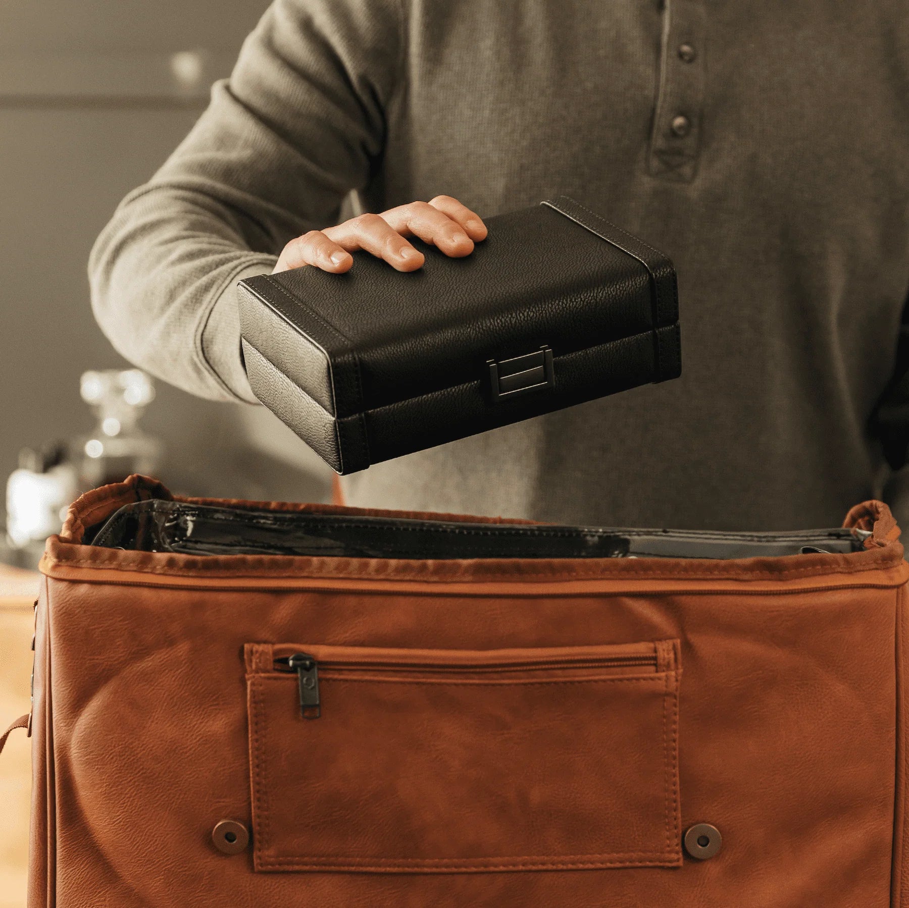 Person holding a black leather cigar case above an open brown leather bag.