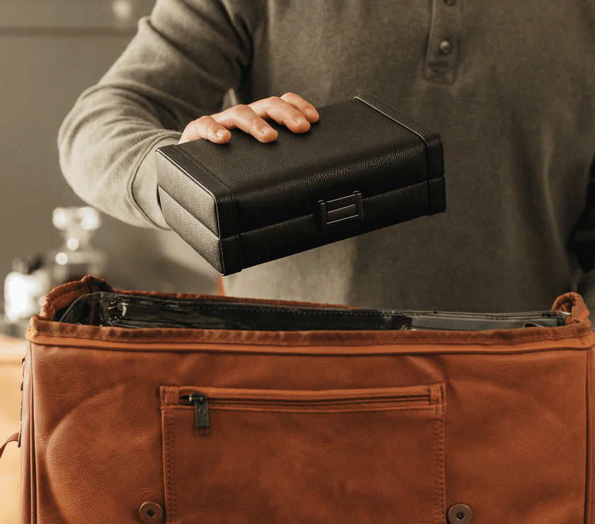 Person holding a black leather cigar case above an open brown leather bag.