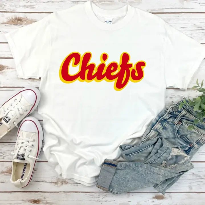 A white short-sleeved t-shirt with the text 'Chiefs' in red and yellow on it, placed on a wooden surface with a pair of sneakers and blue jeans to the side.