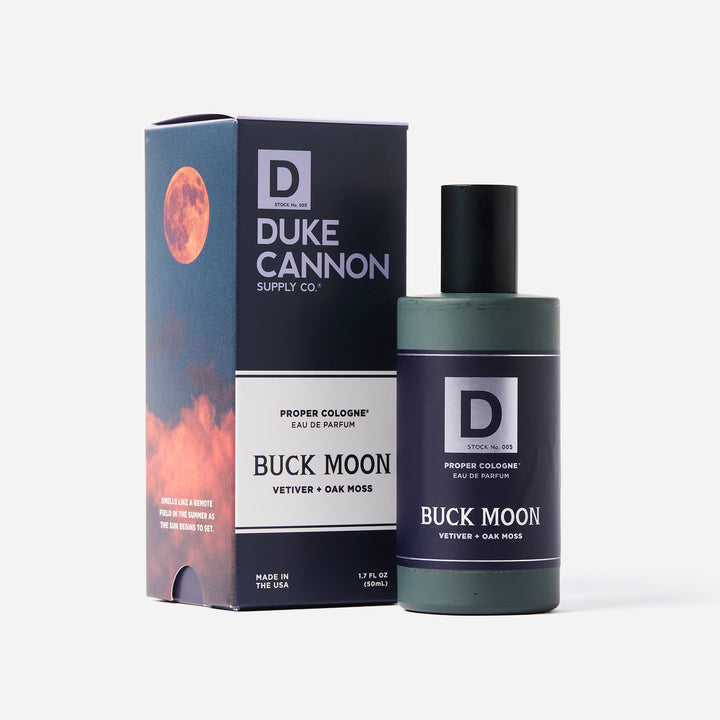 A product image featuring a bottle of Duke Cannon Proper Cologne in the scent 'Buck Moon', alongside its packaging box. The box displays a moon and sunset graphic.
