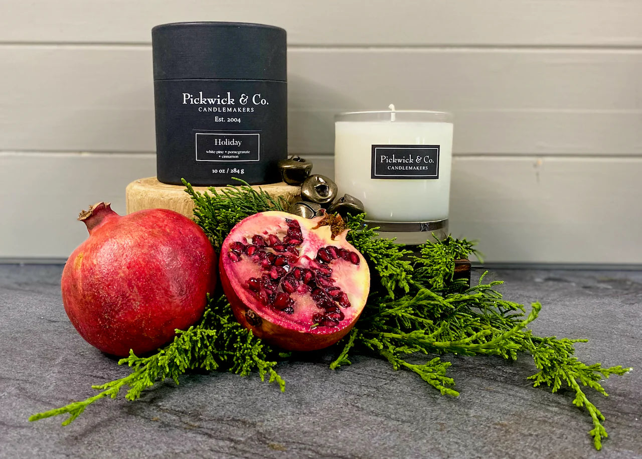 Two candles from Pickwick & Co. with a pomegranate and greenery on a gray surface.
