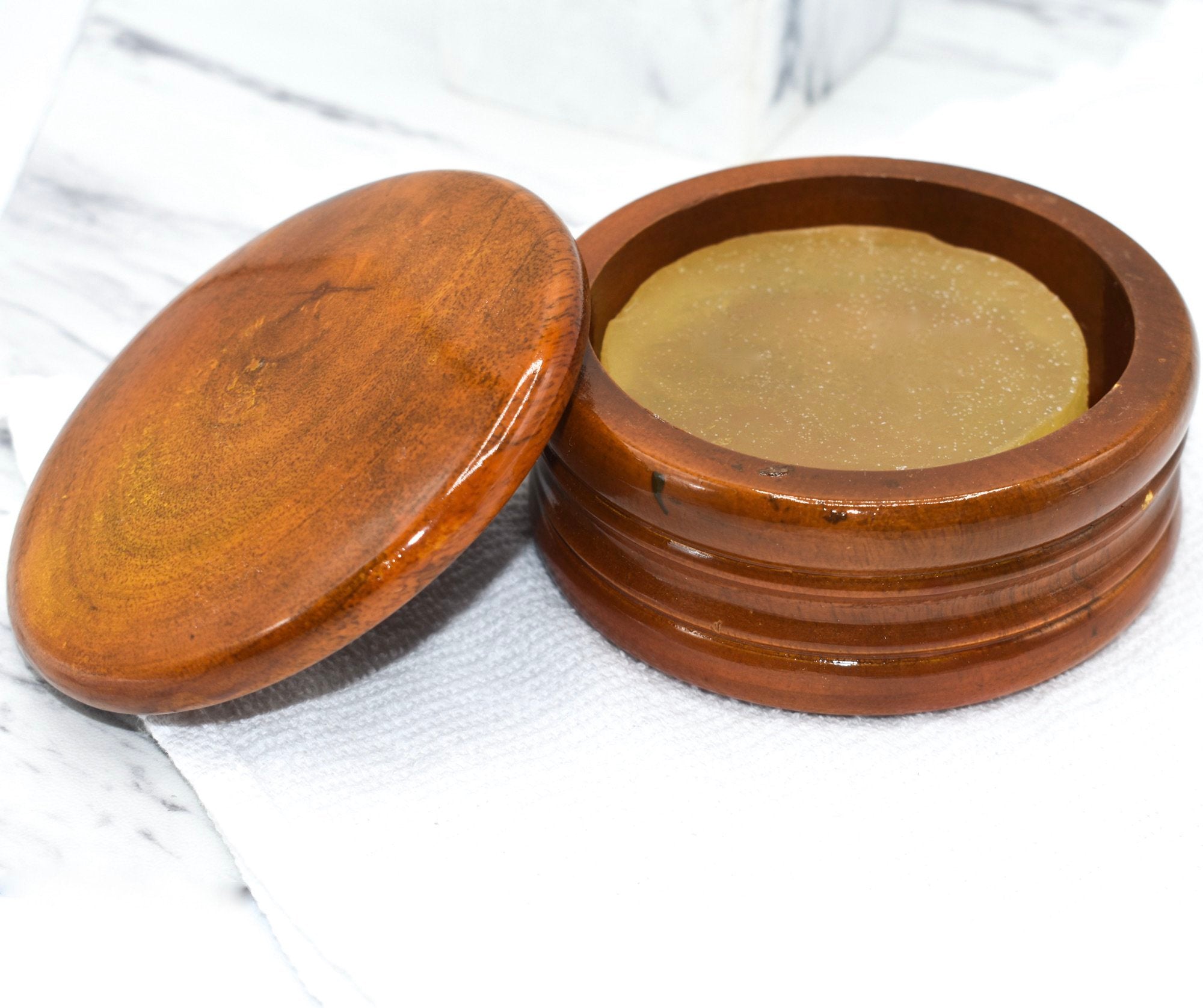 A honey colored mango wood shaving bowl with shave soap inside, sitting on top of a white washcloth and marble countertop.