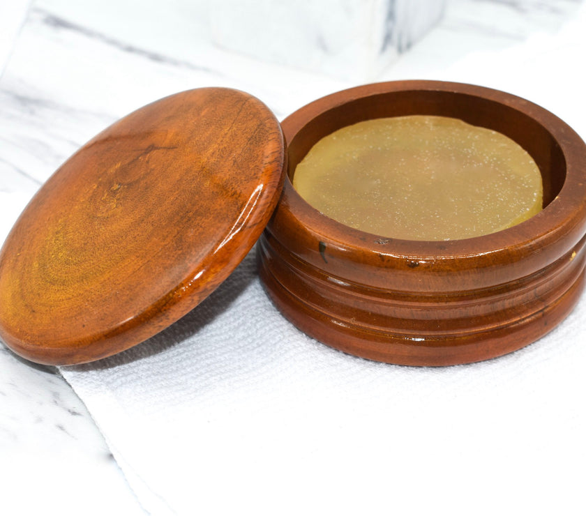 A honey colored mango wood shaving bowl with shave soap inside, sitting on top of a white washcloth and marble countertop.