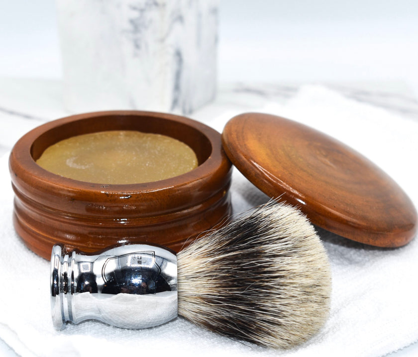 A honey mango wood shaving bowl with the lid off to show shave soap inside, next to a shaving brush and on top of a white washcloth.