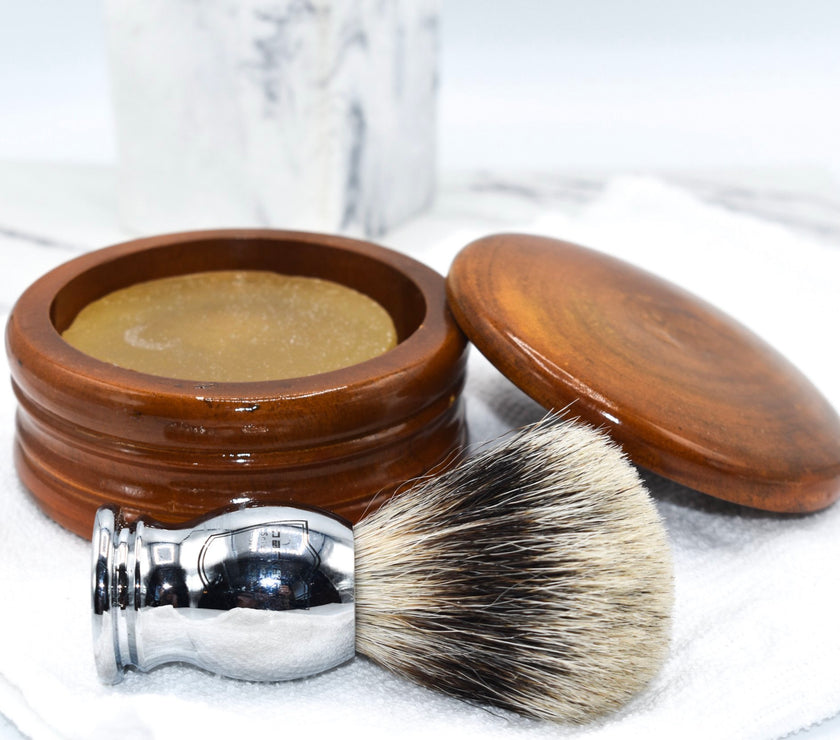 A honey mango wood shaving bowl with the lid off to show shave soap inside, next to a shaving brush and on top of a white washcloth.