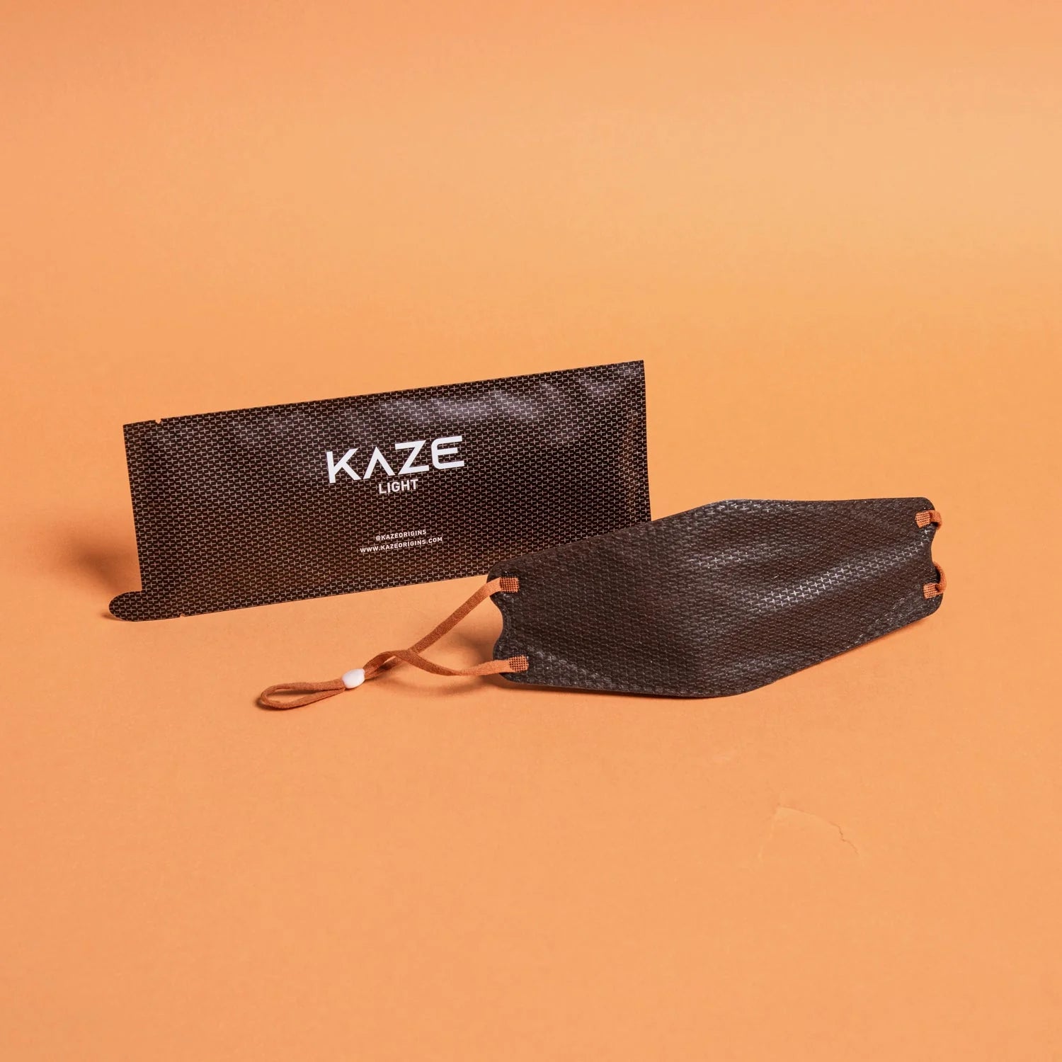 Black mesh face mask with brown strap on an orange background, featuring the brand 'Kaze'.