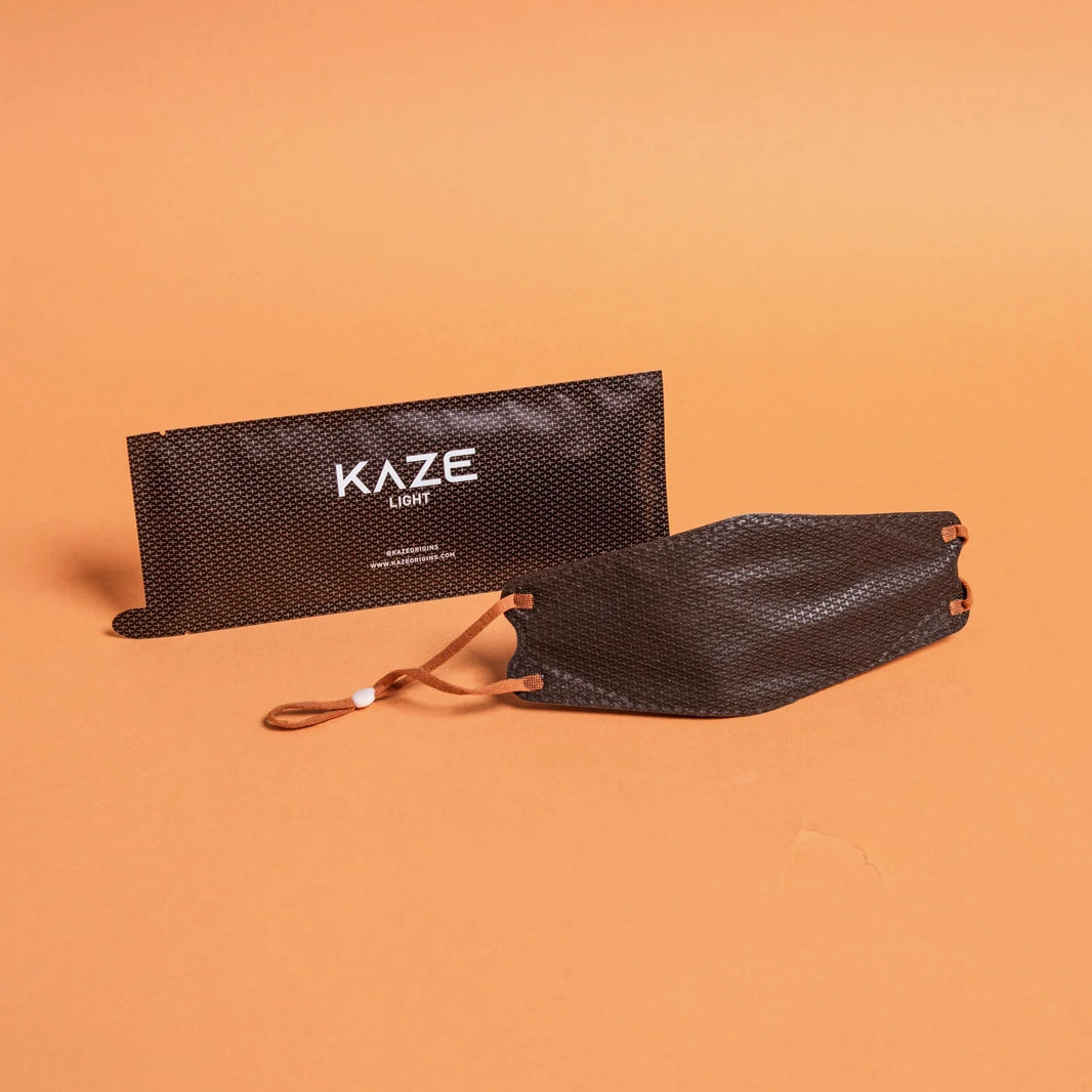 Black mesh face mask with brown strap on an orange background, featuring the brand 'Kaze'.