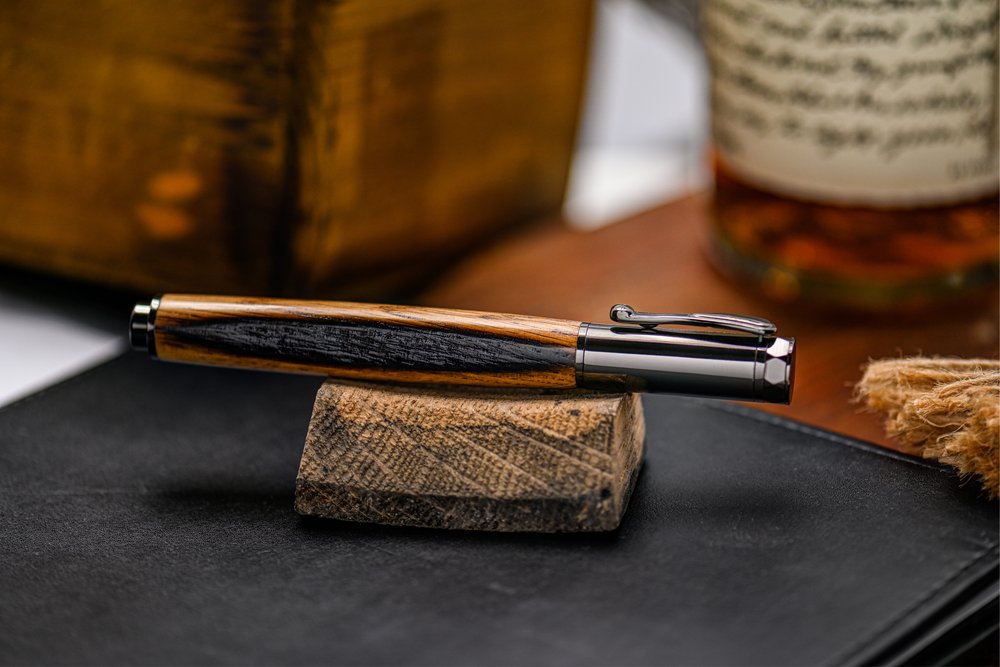 A wood-styled pen with metal cap and clip setting on a small wooden piece on a black folder near a bottle and wooden box.