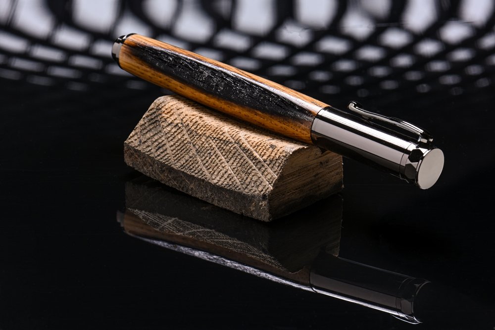 A wooden-styled pen with metal cap and clip resting on a small wood piece on a reflective black surface.
