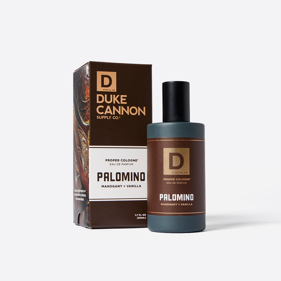 Packaging and bottle of Duke Cannon Proper Cologne in the scent 'Palomino'. The product is packaged in a brown box with the brand and product name, and a bottle with a dark cap and label displaying the same branding and scent name.