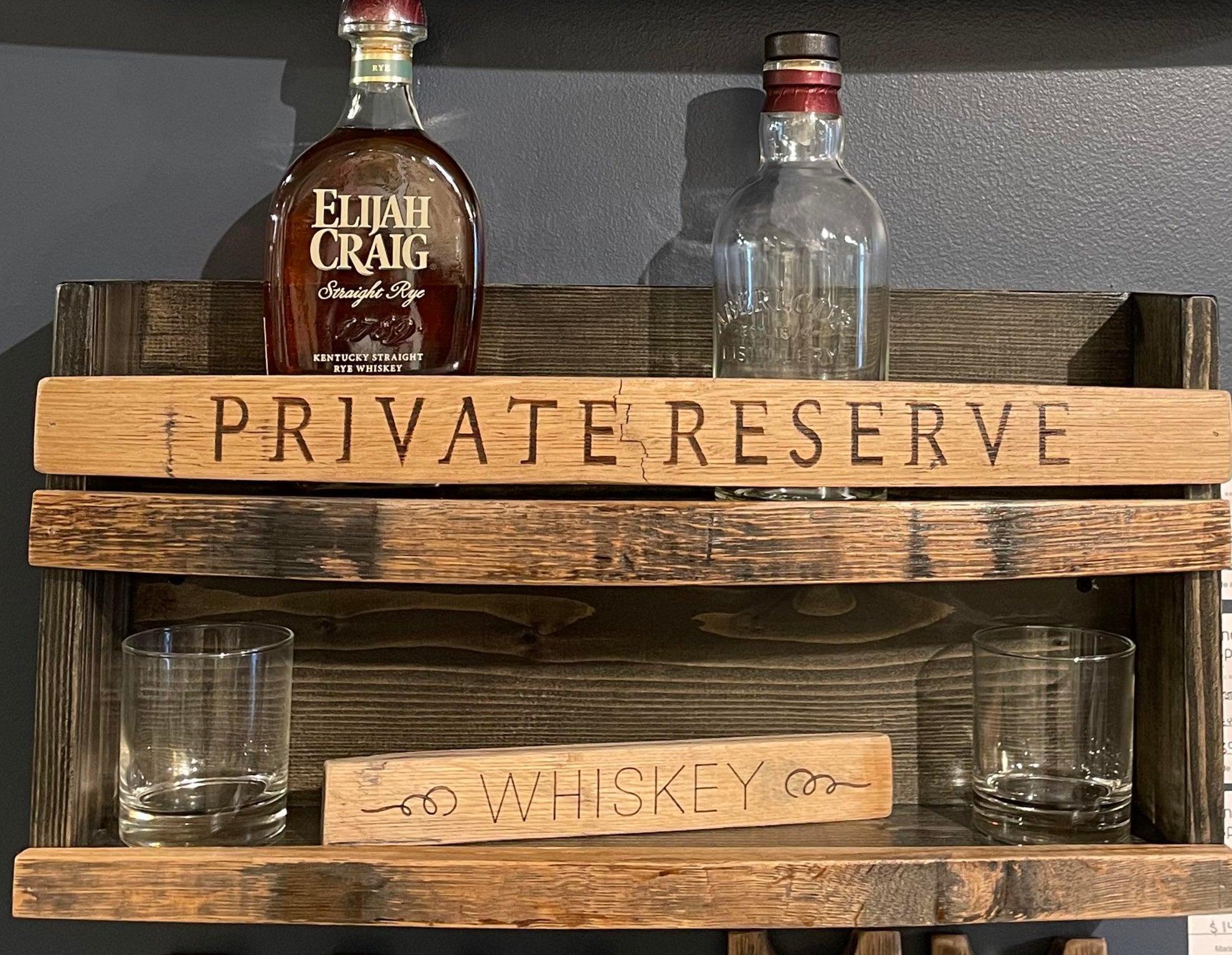 Wooden whiskey rack with 'Private Reserve' and 'Whiskey' signs, Elijah Craig bottle, and glasses.
