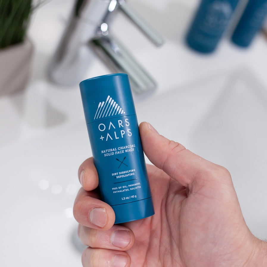 A person holding a container of Oars + Alps Exfoliating Charcoal Solid Face Wash.
