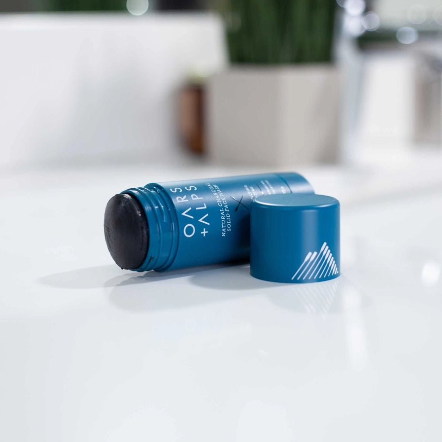 An open container of Oars + Alps Exfoliating Charcoal Solid Face Wash lying on its side on a white surface with the blue cap sitting beside it.