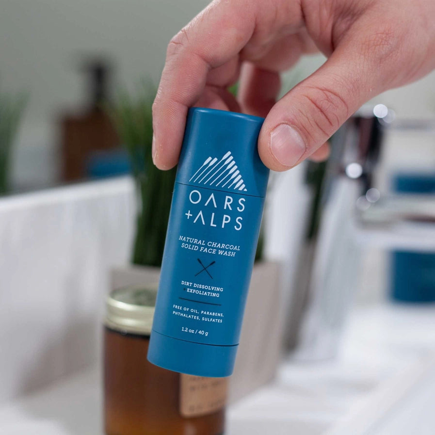 A person holding a container of Oars + Alps Exfoliating Charcoal Solid Face Wash.