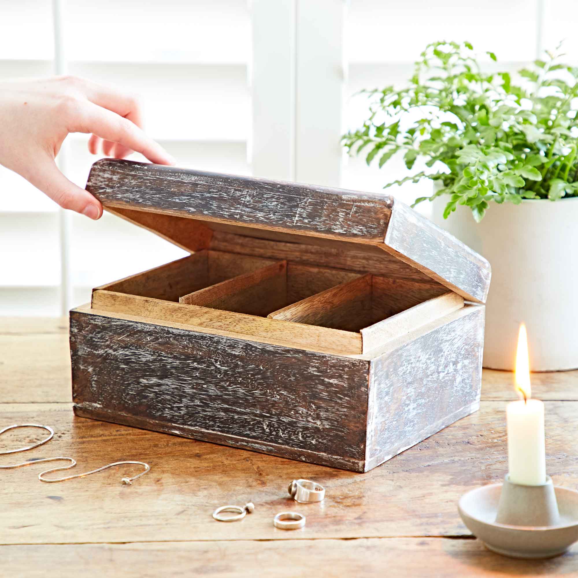 Wooden jewelry box on a wooden surface with a hand reaching towards it, candle, and plant in the background.