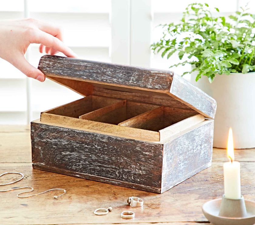 Wooden jewelry box on a wooden surface with a hand reaching towards it, candle, and plant in the background.