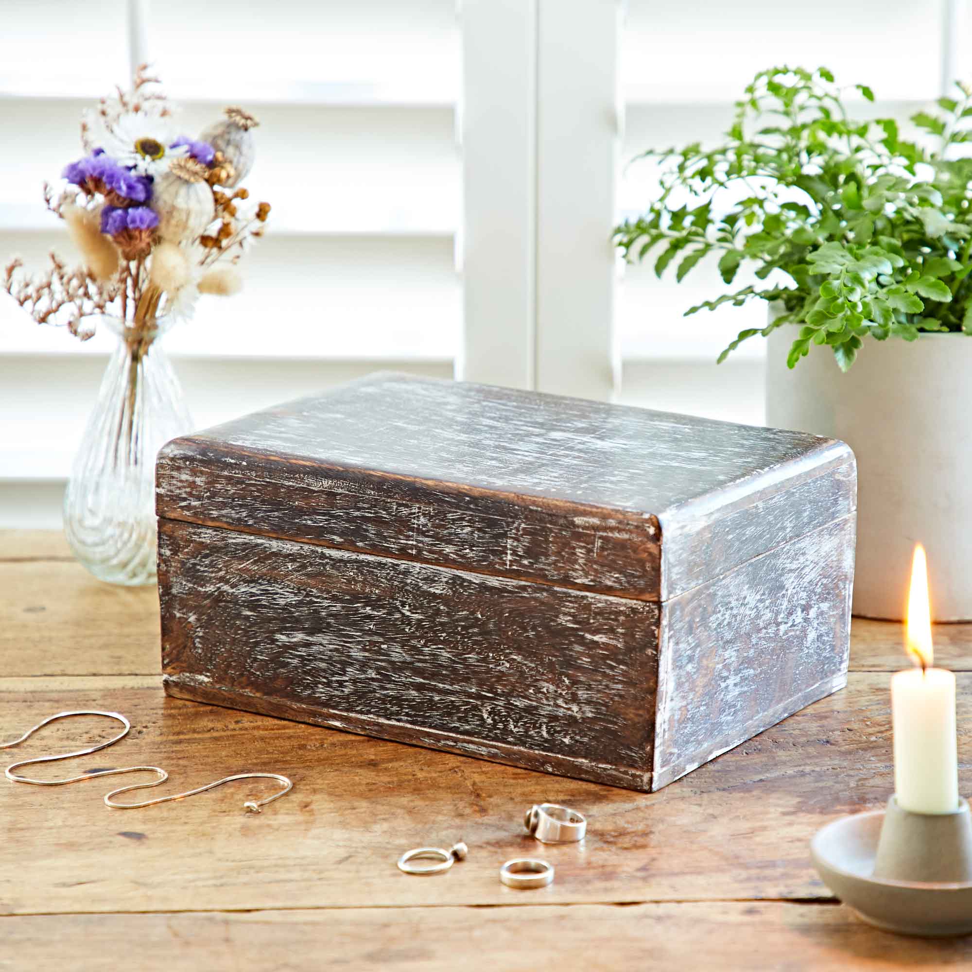 Wooden jewelry box on a wooden surface with rings, a candle, and plants in the background.