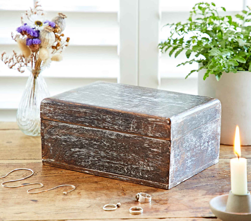 Wooden jewelry box on a wooden surface with rings, a candle, and plants in the background.