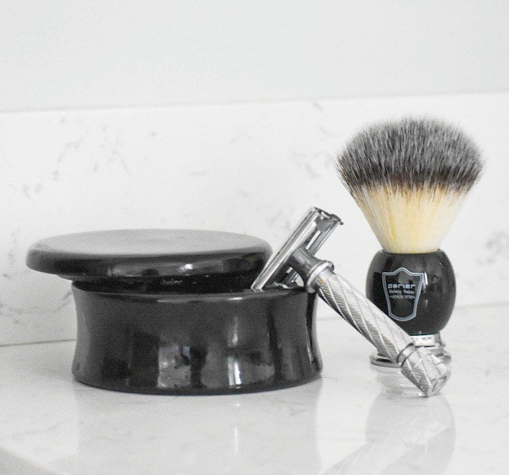 A black mango wood shave bowl with a safety razor leaning against it with a shave brush set next to both on a marble countertop.
