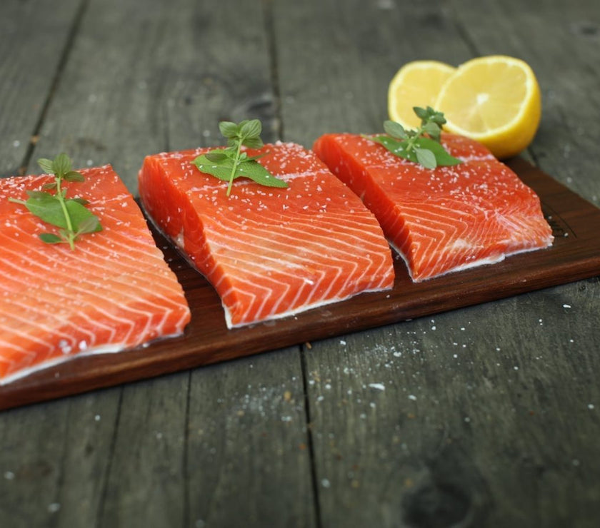 Three pieces of salmon on a wooden grilling board with lemon slices on a dark wooden surface