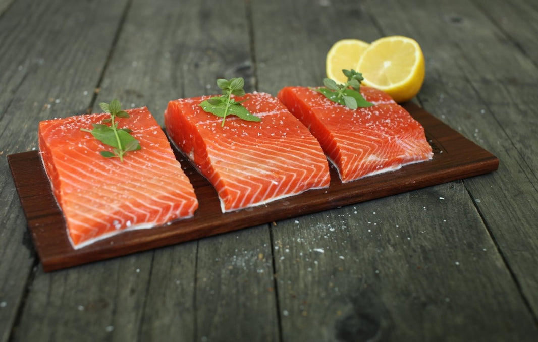Three pieces of salmon on a wooden grilling board with lemon slices on a dark wooden surface