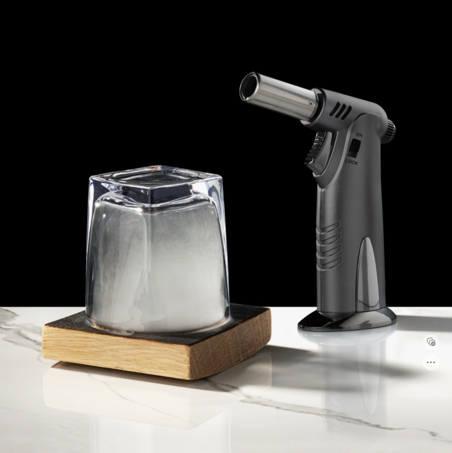 The rocks glass face down on a smoking board with smoke inside, and a butane torch setting beside them.