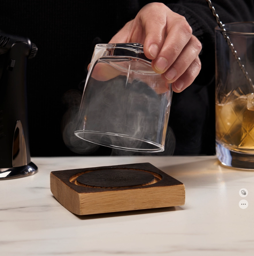 A rocks glass with smoke in it held over a  wood smoking barrel square by a person.