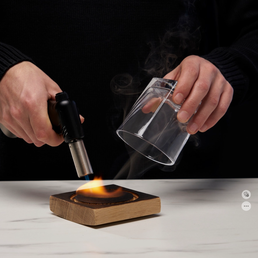 A person using the butane torch on the wood smoking barrel square to create smoke, and the rocks glass held over it in their other hand.