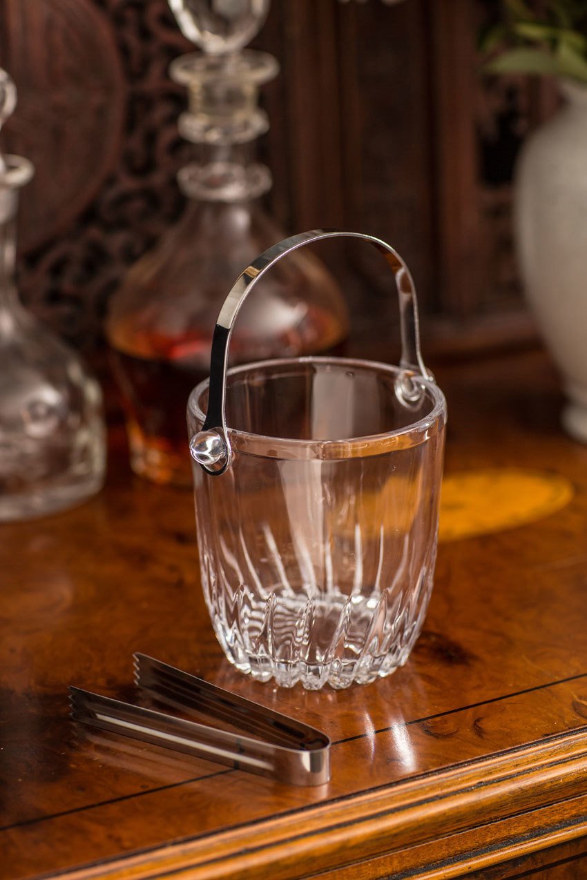 Clear glass ice bucket with silver handle and tongs on a wooden surface