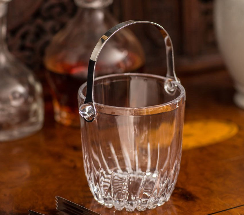 Clear glass ice bucket with silver handle and tongs on a wooden surface
