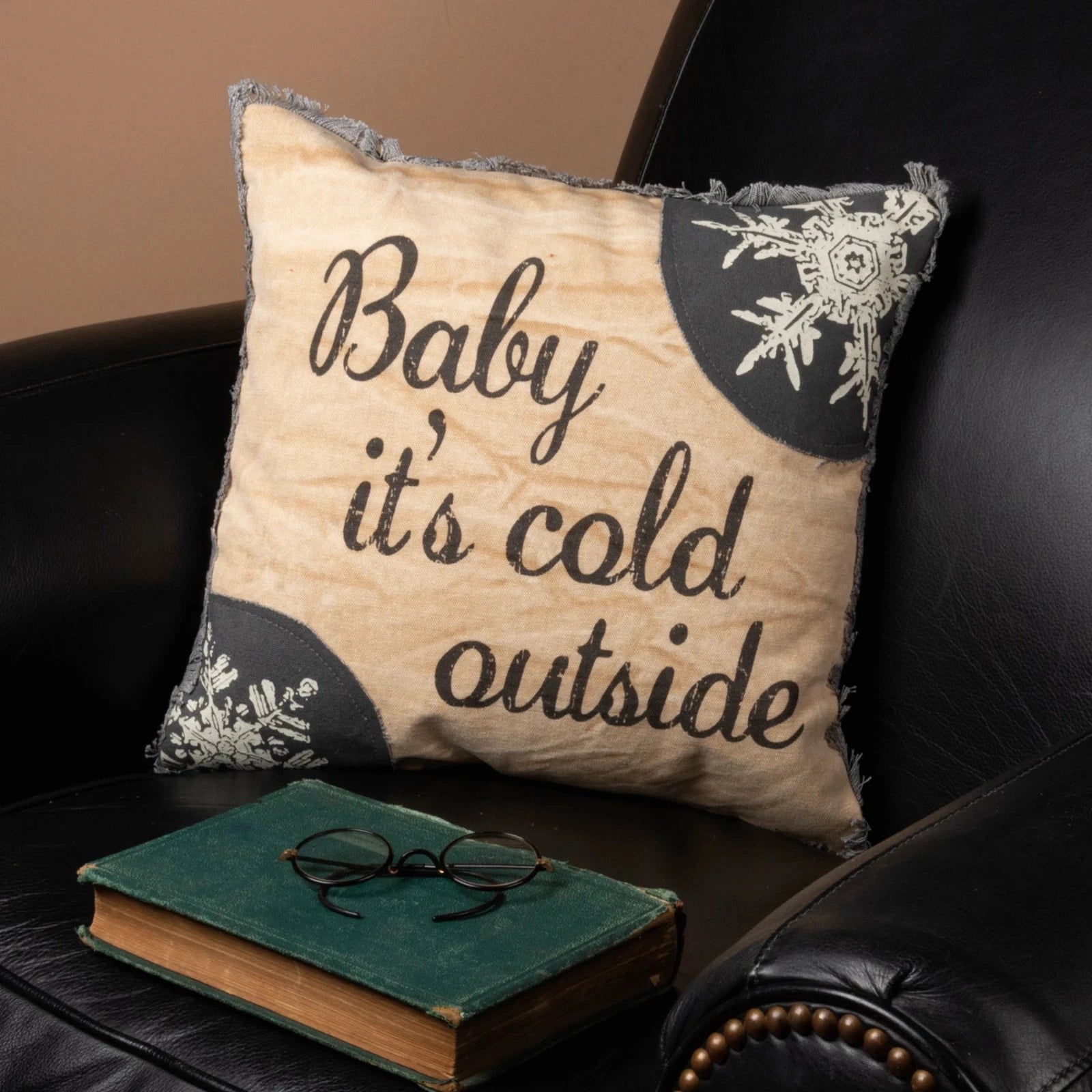 Decorative pillow with 'Baby it's cold outside' text on a black leather couch with a book and glasses.