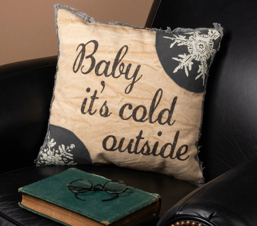Decorative pillow with 'Baby it's cold outside' text on a black leather couch with a book and glasses.