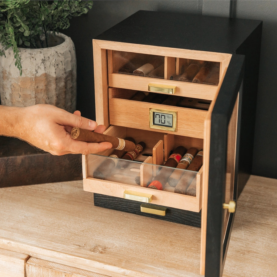 A black ash wood humidor cabinet with gold-plated hardware, featuring a digital display and multiple cigar drawers filled with cigars. A person's hand is adding a cigar to one of the drawers.