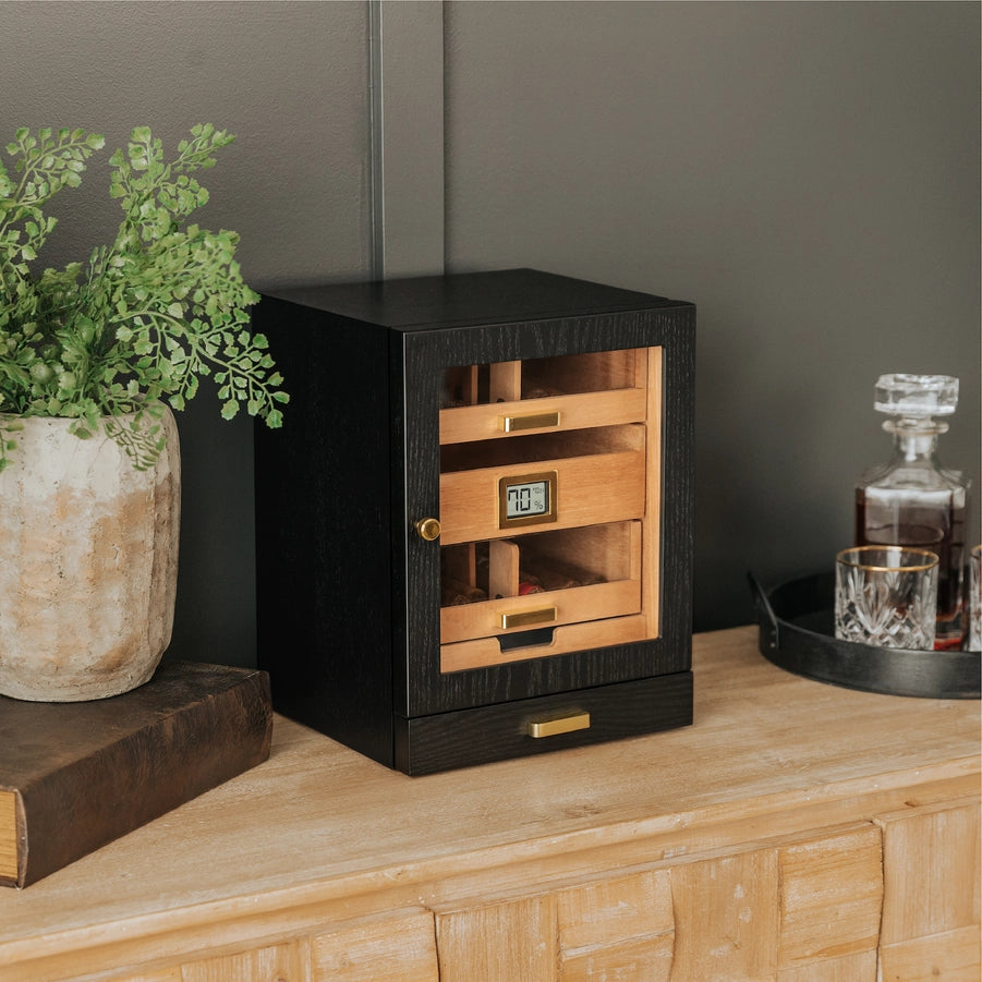 A black ash wood humidor cabinet with gold-plated hardware, featuring a digital display and multiple cigar drawers sitting on a table next to a book and potted plant, and whiskey with glasses.